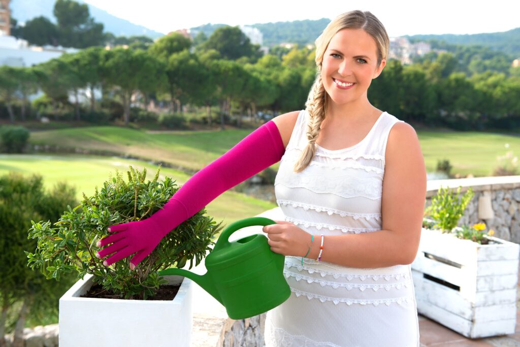 Dicke Beine oder Arme? So erkennen Sie Lipödem & Lymphödem - Ihr Sanitätshaus Kurda in Kiel hilft 1 Frau in weißem Kleid gießt auf einer Terrasse eine Pflanze und trägt dabei einen pinken Kompressionsarmstrumpf mit Handschuh. Erhältlich im Sanitätshaus Kurda in Kiel.