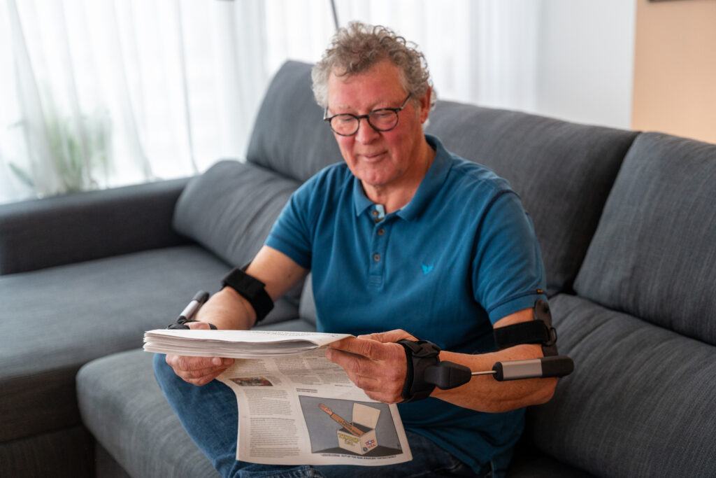 Dämpfen statt Fixieren: So funktioniert die Stil Hand Control™ bei Tremor 1 Mann sitzt auf dem Sofa und liest mithilfe der Stil Hand Control - Stilorthese Zeitung.