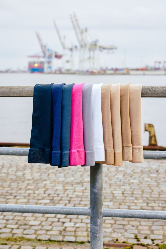 Verschiedenfarbige Kompressionsstrümpfe hängen vor einem Hafen an einem Geländer.