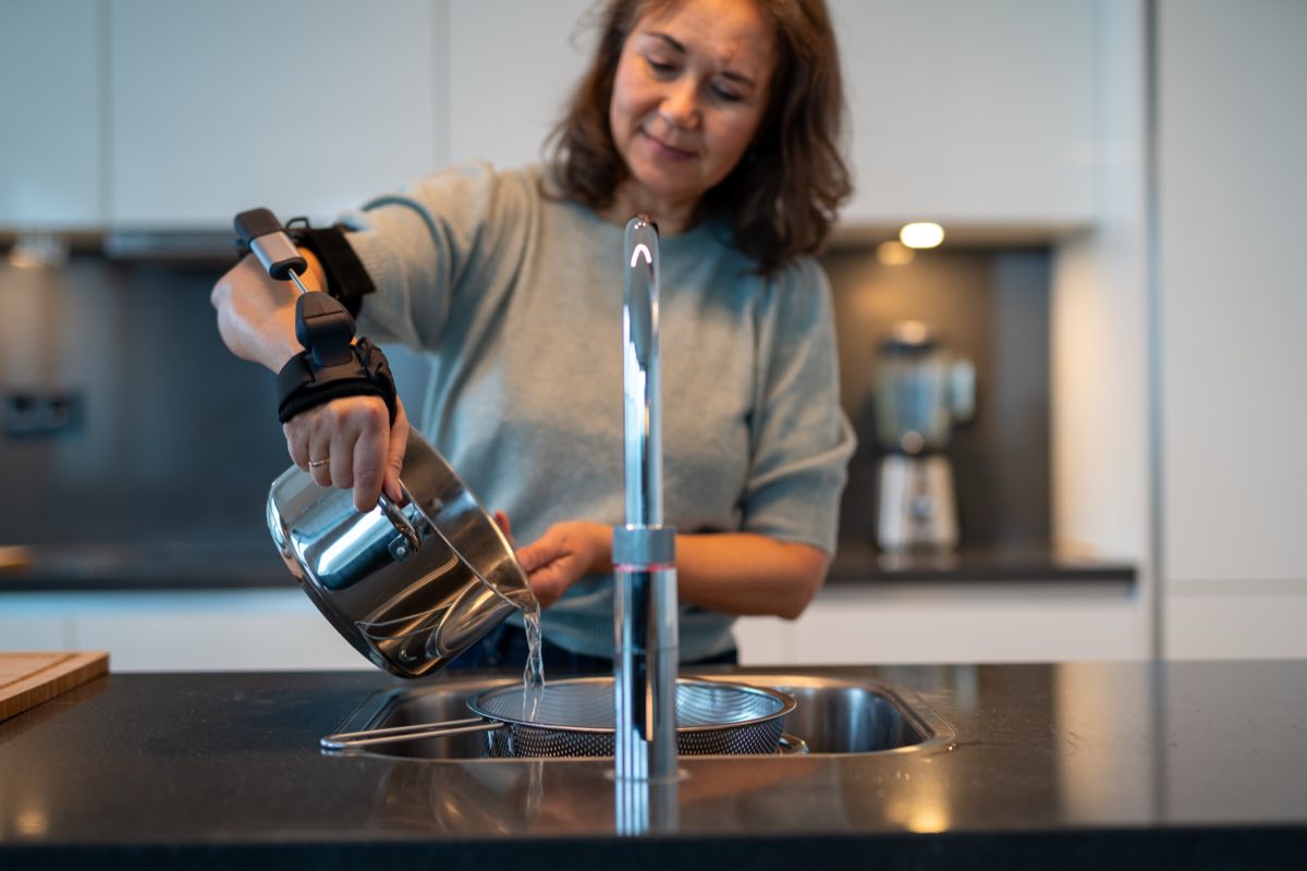 Frau mit Stilorthese am Unterarm gießt konzentriert Wasser aus einem Topf ab