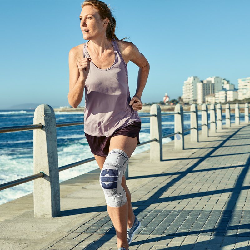 Frau joggt auf einem Steg am Meer, am Knie trägt sie eine stabilisierende Knieorthese