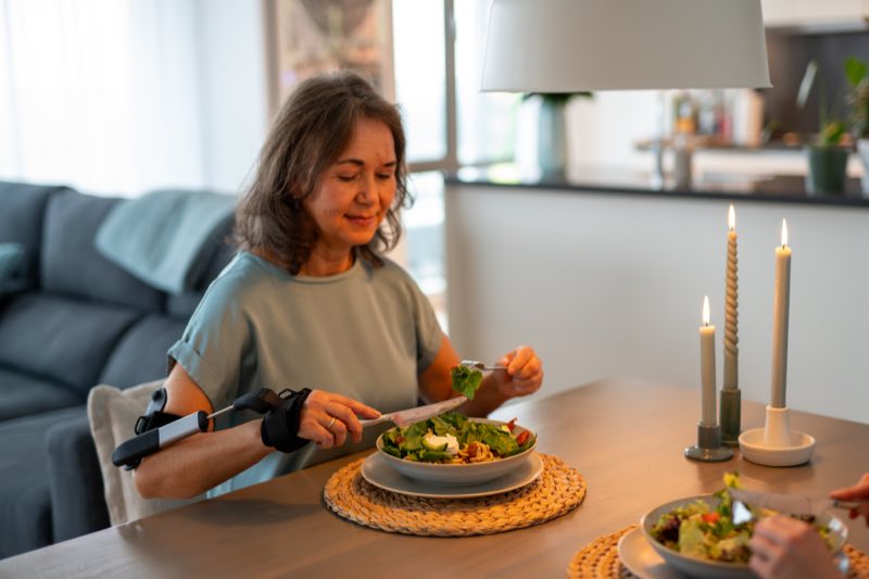 Frau sitzt entspannt am gedeckten Tisch und isst, am Unterarm trägt sie die Stilorthese Stil Hand Control™