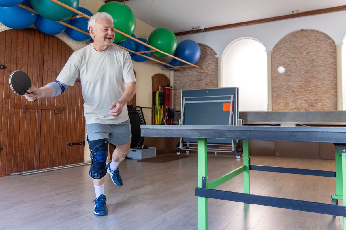 Älterer Mann spielt lachend Tischtennis, am Knie trägt er eine stabilisierende Orthese