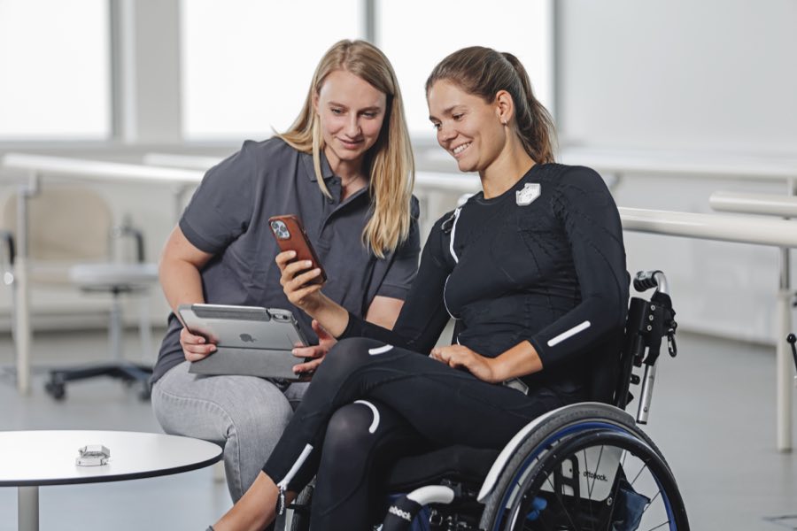 Rollstuhlnutzerin im Exopulse Suit im Gespräch mit einer Fachkraft, beide schauen gemeinsam auf ein Tablet
